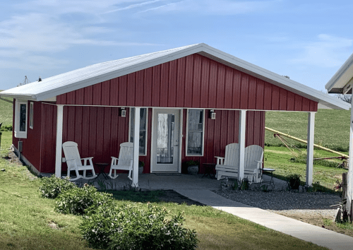 A red cabin with a white roof, featuring two rocking chairs on the porch and a grassy area in front.