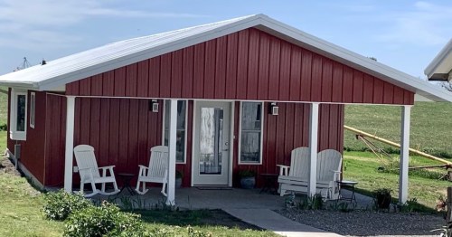 A red farmhouse with a white roof, featuring a porch and two white chairs, surrounded by green grass and plants.