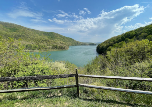 A scenic view of a river winding through green hills under a blue sky with fluffy clouds. A wooden fence in the foreground.