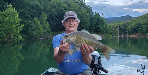 A smiling man in a boat holds a large fish, surrounded by lush green trees and calm water.