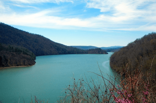 A serene lake surrounded by mountains and trees under a blue sky with wispy clouds.