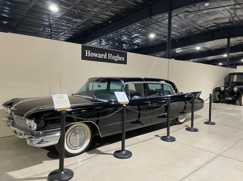 A vintage black limousine displayed in a museum, labeled "Howard Hughes," with informational plaques and barriers.