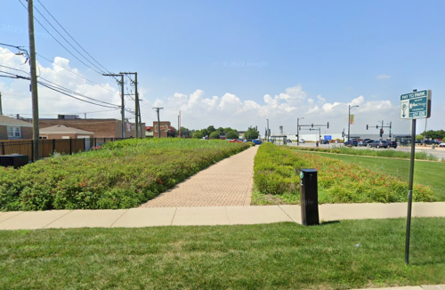 A grassy area with a paved path, power lines, and a street sign in a suburban setting under a clear blue sky.