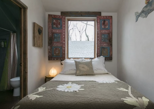 Cozy bedroom with a bed, decorative window shutters, and a view of trees outside. A bathroom is visible in the background.