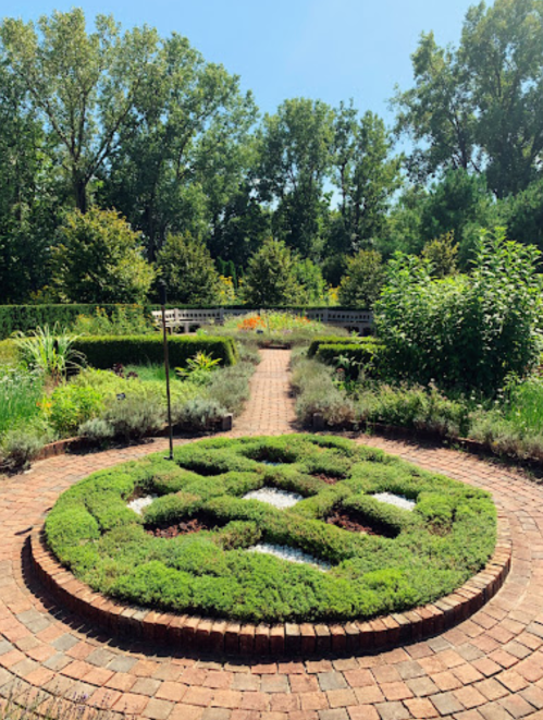 A circular garden design with neatly trimmed greenery and a brick pathway, surrounded by trees and bright blue sky.