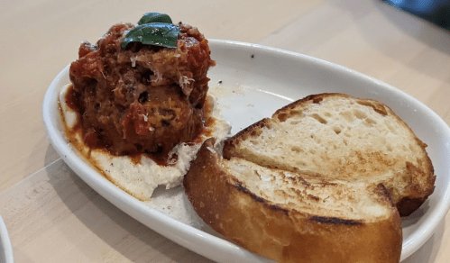 A meatball topped with marinara sauce and basil, served with two slices of toasted bread on a white plate.