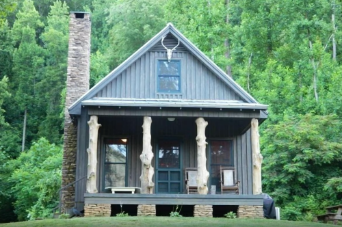 A rustic wooden cabin with a stone chimney, surrounded by lush green trees and featuring unique log pillars on the porch.