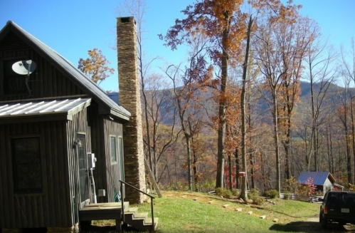 A rustic cabin with a stone chimney surrounded by autumn trees and mountains in the background.