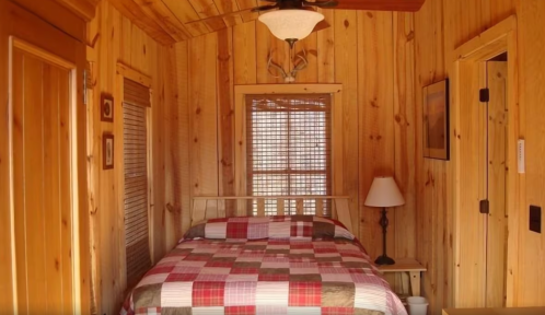Cozy wooden bedroom with a plaid quilt, lamp, and rustic decor, featuring a window with blinds and a ceiling fan.
