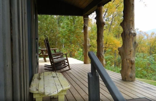A rustic porch with rocking chairs and a wooden bench, surrounded by trees in autumn colors.
