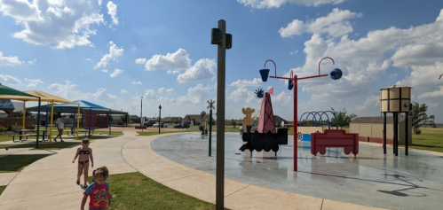 A sunny park scene with children playing near a colorful water play area and shaded picnic spots.
