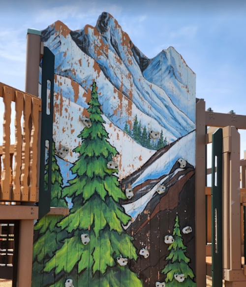Colorful climbing wall with a mountain and pine tree mural, part of a playground structure.