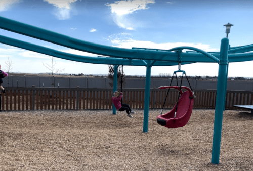 A child swings on a playground swing set under a blue sky, with a second swing visible in the background.