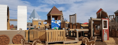 Colorful wooden playground with various structures, including slides and climbing areas, under a blue sky.