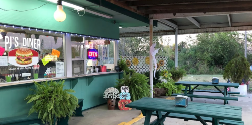 Outdoor seating area of PJ's Diner, featuring green picnic tables, plants, and an open sign in the window.