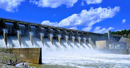A large dam with water flowing from multiple gates, under a blue sky with fluffy clouds, and people standing nearby.