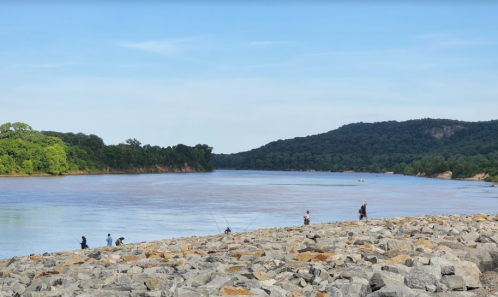People fishing along a rocky riverbank with lush greenery and hills in the background under a clear blue sky.