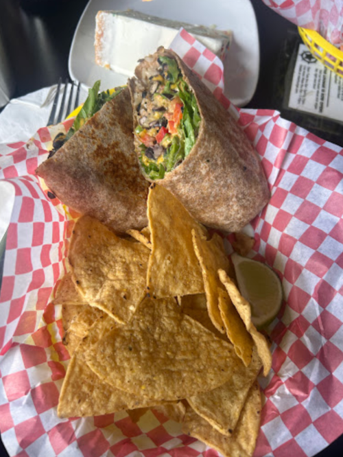 A whole grain wrap filled with vegetables and beans, served with tortilla chips and a lime wedge in a red-checkered basket.
