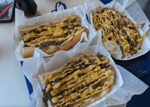 Three sandwiches filled with beef and topped with melted cheese sauce, served on a tray with a drink in the background.