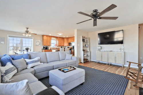Bright living room with a gray sectional sofa, coffee table, and a TV, featuring wooden floors and a ceiling fan.