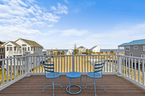A balcony view with two blue chairs and a small table overlooking houses and a clear sky.
