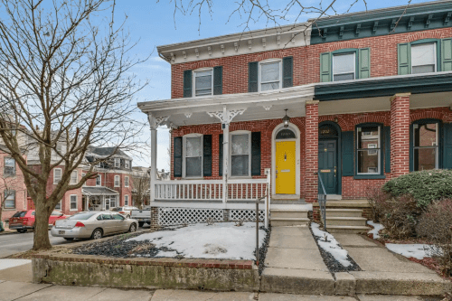 A brick townhouse with a yellow door, porch, and snow on the ground, surrounded by other homes on a quiet street.