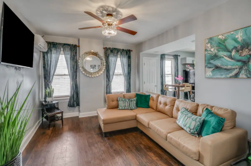 Cozy living room with a tan sectional sofa, decorative pillows, a ceiling fan, and large windows with curtains.