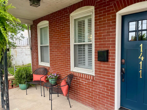 A brick house entrance with two chairs, potted plants, and a blue front door displaying the number 1411.