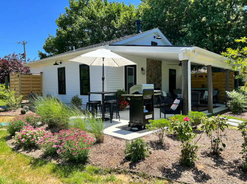 A cozy house with a patio, grill, and garden, surrounded by greenery and flowers on a sunny day.