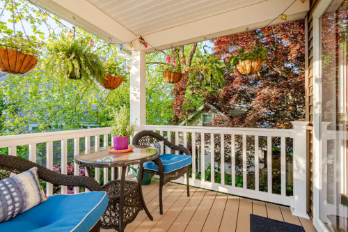 A cozy porch with two blue chairs, a small table, and hanging plants, surrounded by vibrant greenery.