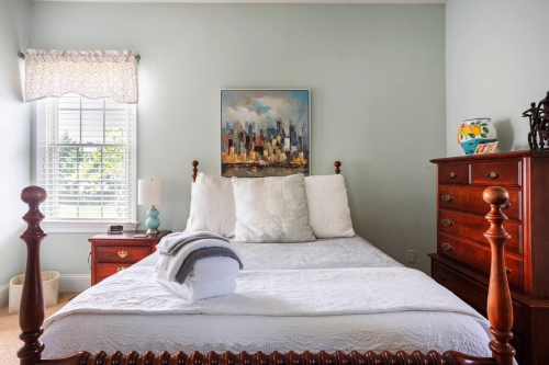A cozy bedroom featuring a wooden bed, decorative pillows, a dresser, and a cityscape painting on the wall.