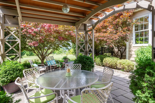 A cozy outdoor patio with a round table, chairs, and lush greenery, surrounded by colorful trees and a sunny view.