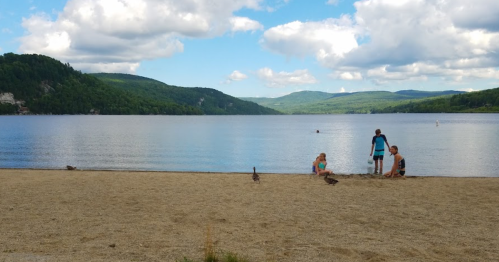 A sandy beach by a calm lake, with people playing and mountains in the background under a partly cloudy sky.