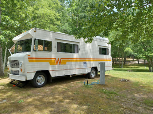 A vintage RV parked on grassy land surrounded by trees, with a water hookup nearby.