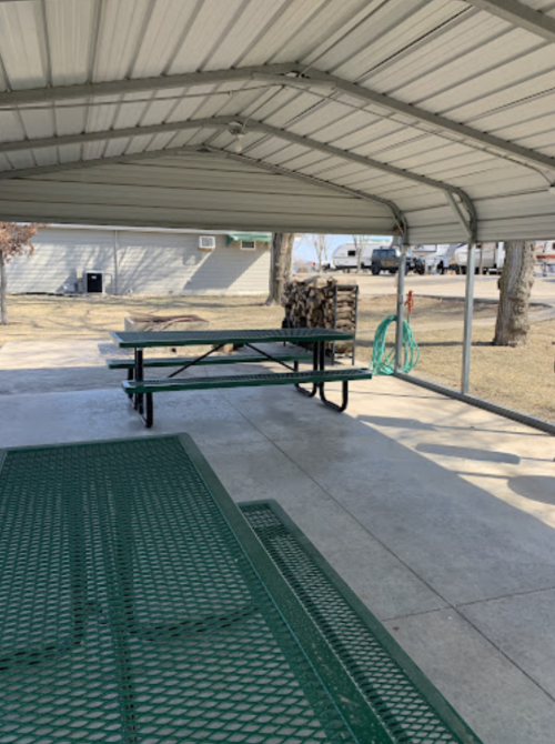 A covered picnic area with a green table and benches, concrete floor, and trees in the background.
