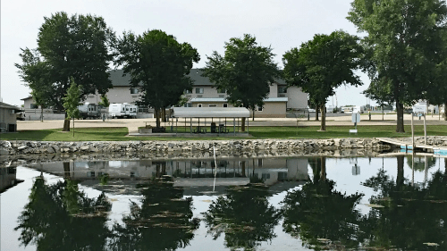 A calm lake reflects trees and a building, with a gazebo on the shore and a grassy area in the background.