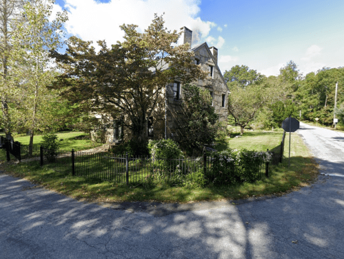A stone house surrounded by greenery at a street corner, with a fence and trees under a clear blue sky.