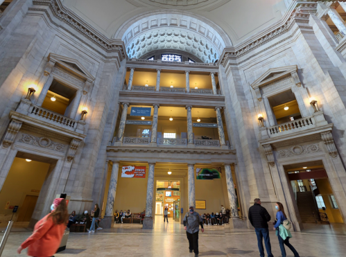 Interior of a grand building with high ceilings, large windows, and people walking through a spacious atrium.