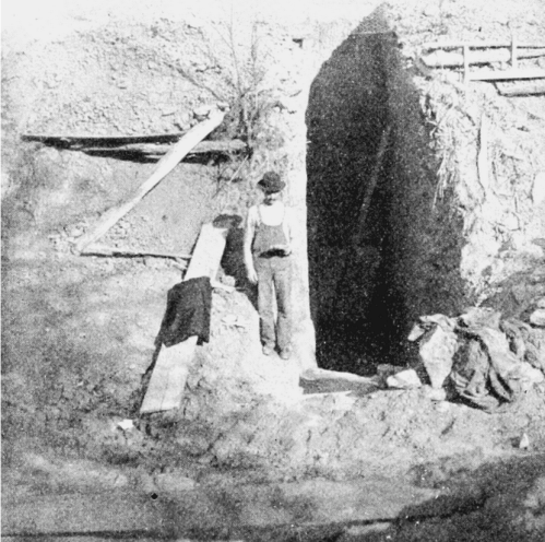A historical black-and-white photo of a man standing at the entrance of a mine, with mining tools and clothing nearby.