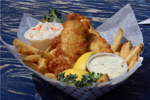A basket of fried fish, fries, coleslaw, and lemon wedges, served with a side of tartar sauce on a blue table.