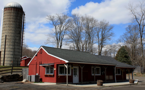 A red barn-style building with a silo nearby, surrounded by trees and a blue sky with clouds.