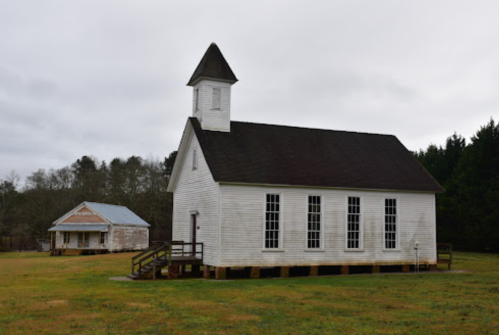 A white, wooden church with a steeple beside an old, weathered house in a grassy field under a cloudy sky.