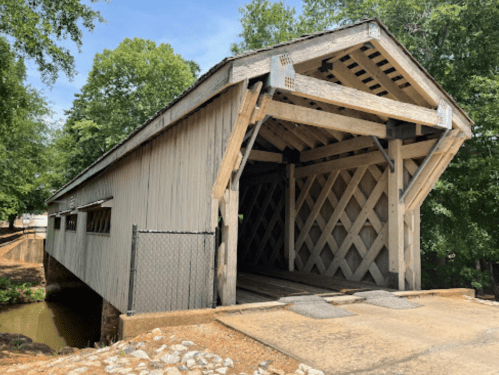 A wooden covered bridge with a lattice design, surrounded by trees and a small creek.