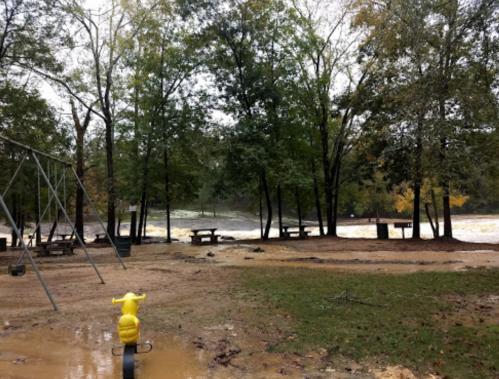 A playground with a yellow tricycle in the foreground, surrounded by trees and a river in the background, under cloudy skies.