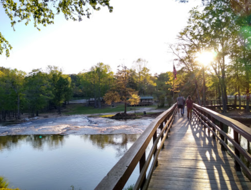 A wooden bridge over a calm river at sunset, with people walking and trees lining the banks.