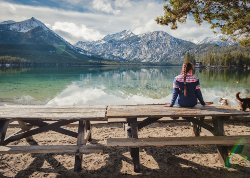 A person sits on a picnic table by a serene lake, surrounded by mountains and trees, reflecting on the water.