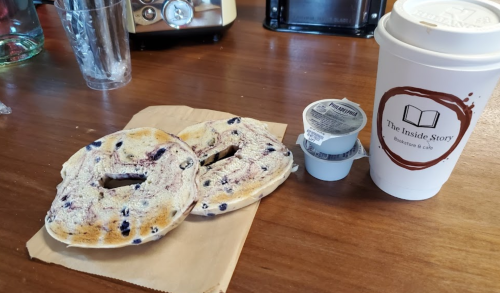 A wooden table with two blueberry bagels, cream cheese, and a coffee cup from The Inside Story bookstore and café.