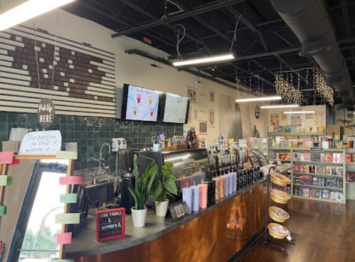 A cozy café interior with a coffee counter, menu screens, plants, and shelves filled with books and snacks.