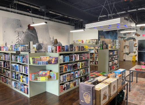 A cozy bookstore with shelves filled with books and games, featuring a mural of mountains on the wall.