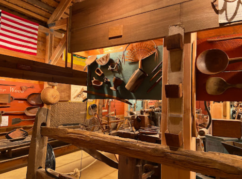 Interior of a woodworking shop displaying various tools and wooden items, with an American flag in the background.